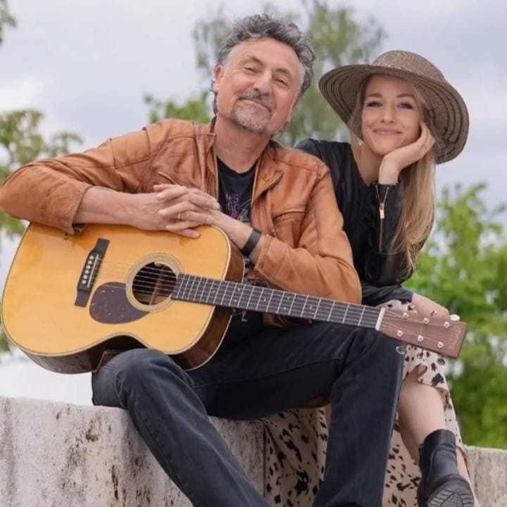 Mann spielt Gitarre, Frau lächelt im Park, Natur im Hintergrund.
